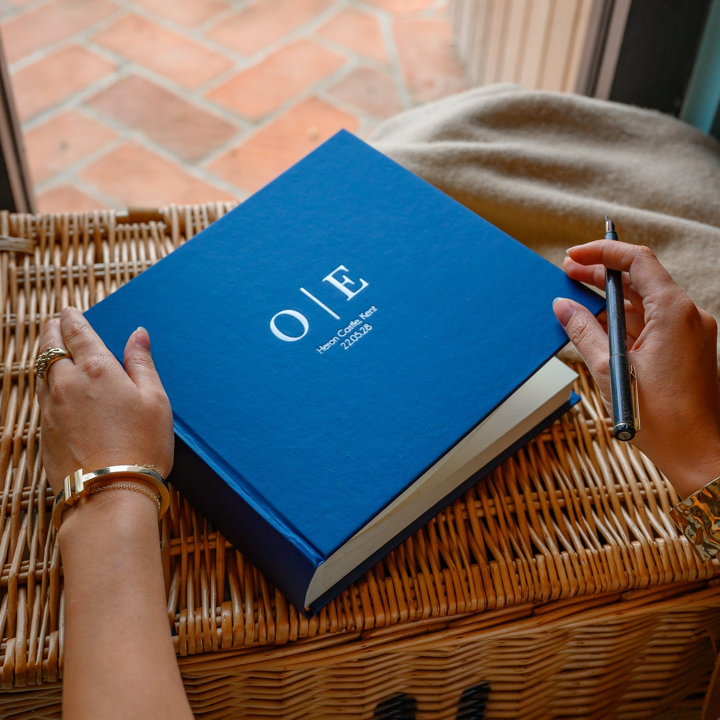 Woman holding a personalised wedding guest book with 'O&E' hand printed on the front, on a wicker surface. 