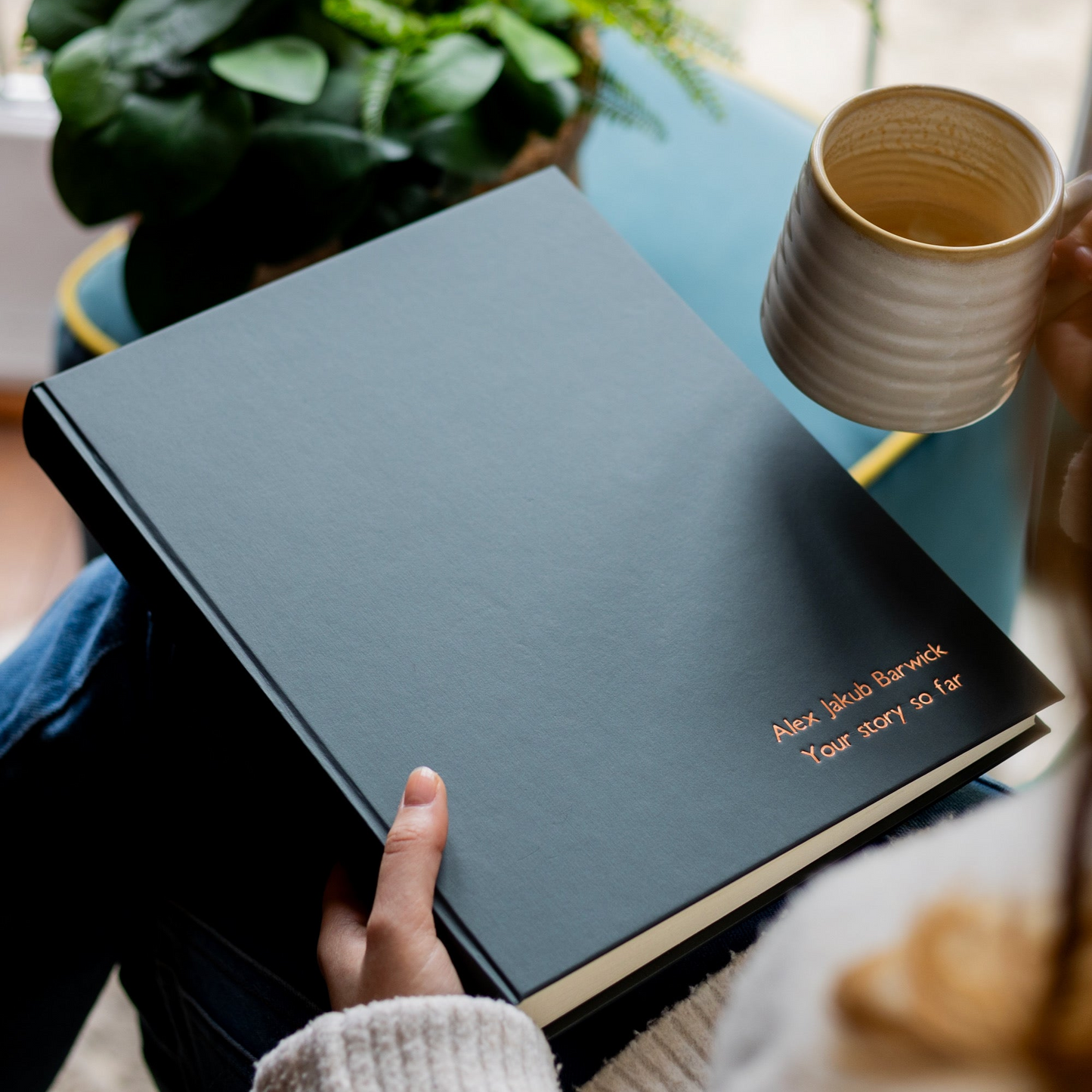 Person holding a black hardback photo album personalised with printed text and a mug on a table