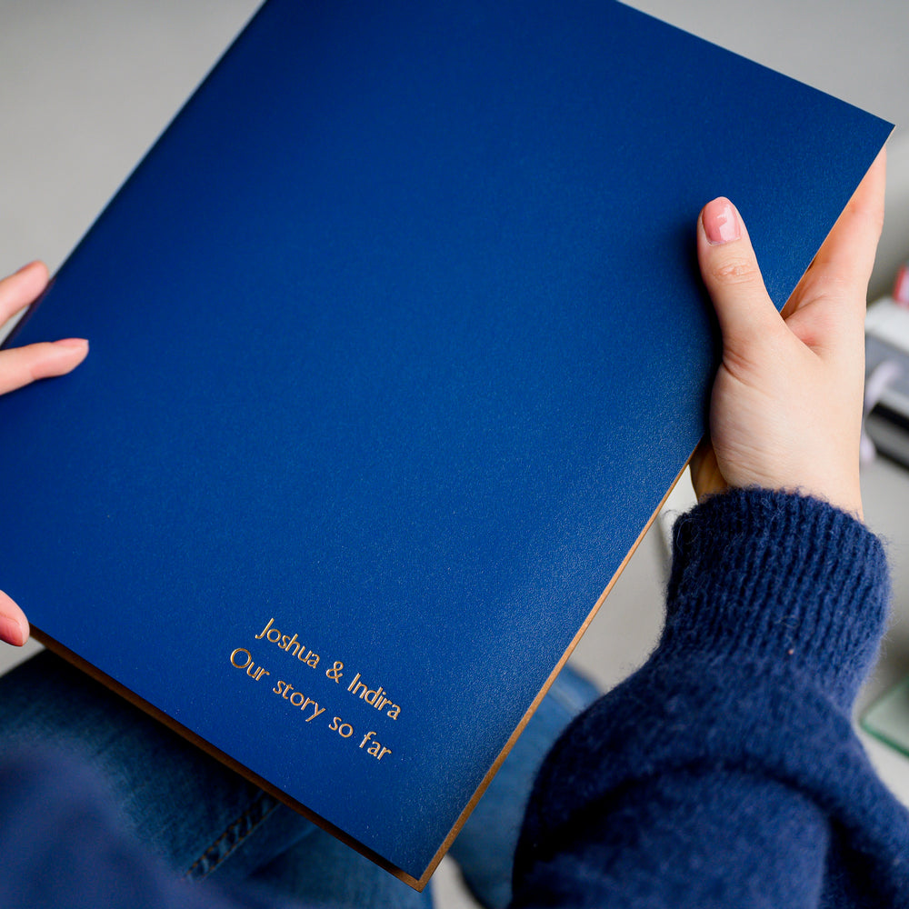 A woman in a blue jumpeer and a pair of jeans is holding a blue A4 photo album in her hands. In the background is a cup of coffee and a pile of books. The A4 album has been personalised with gold writing.