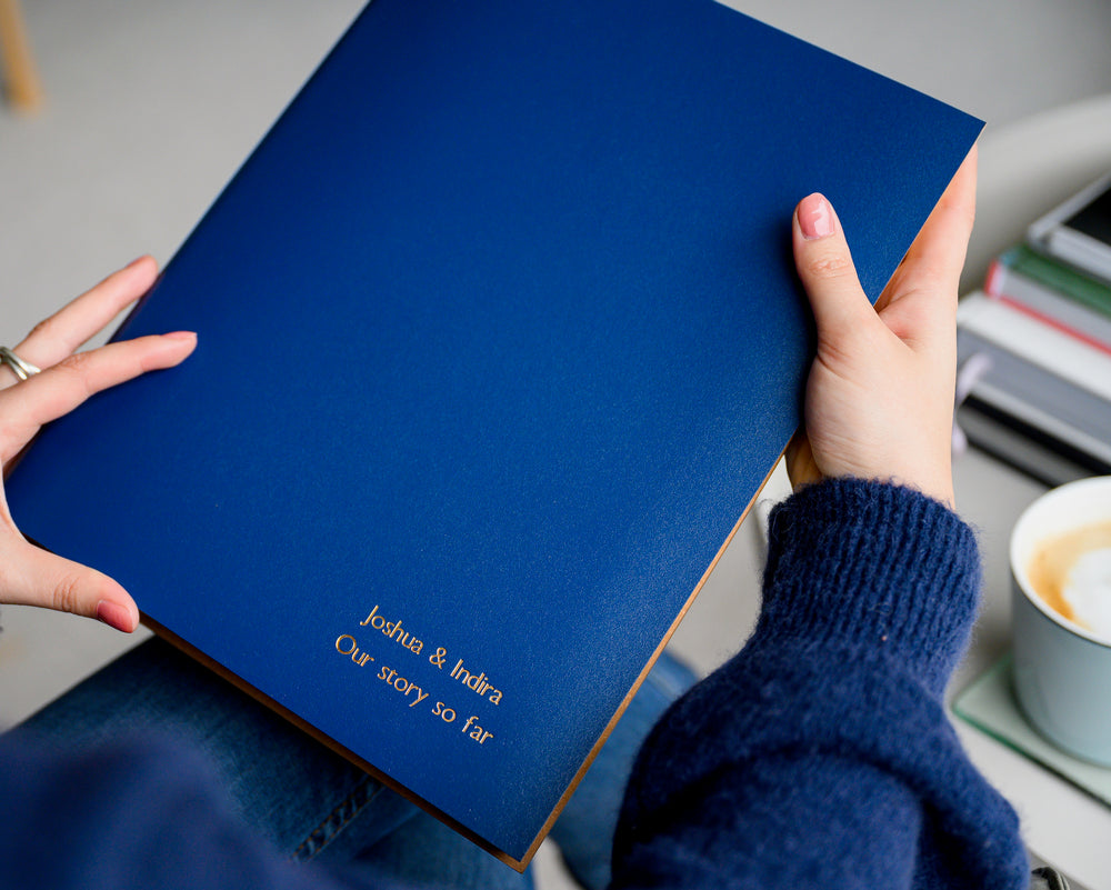 A woman in a blue jumpeer and a pair of jeans is holding a blue A4 photo album in her hands. In the background is a cup of coffee and a pile of books. The A4 album has been personalised with gold writing.