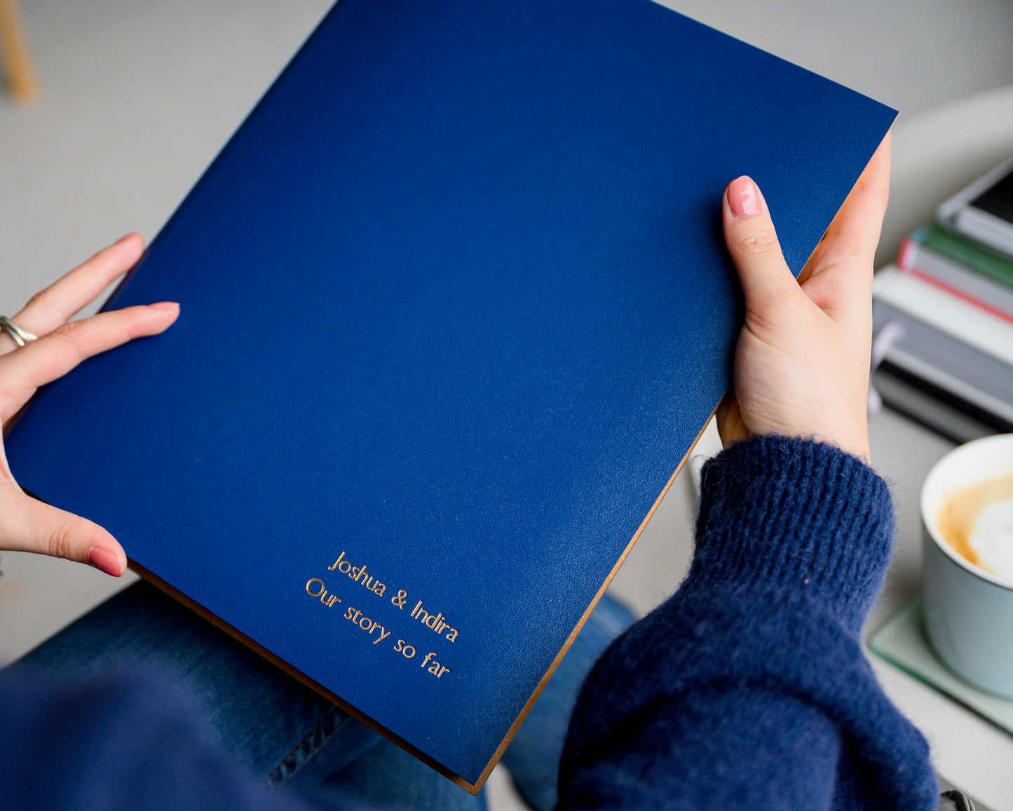 A woman in a blue jumpeer and a pair of jeans is holding a blue A4 photo album in her hands. In the background is a cup of coffee and a pile of books. The A4 album has been personalised with gold writing.