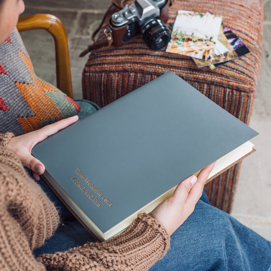 a woman is sitting in a chair with an A4 photo album on her lap. She is wearing a brown jumper and a pair of jeans. There is a camera in the background