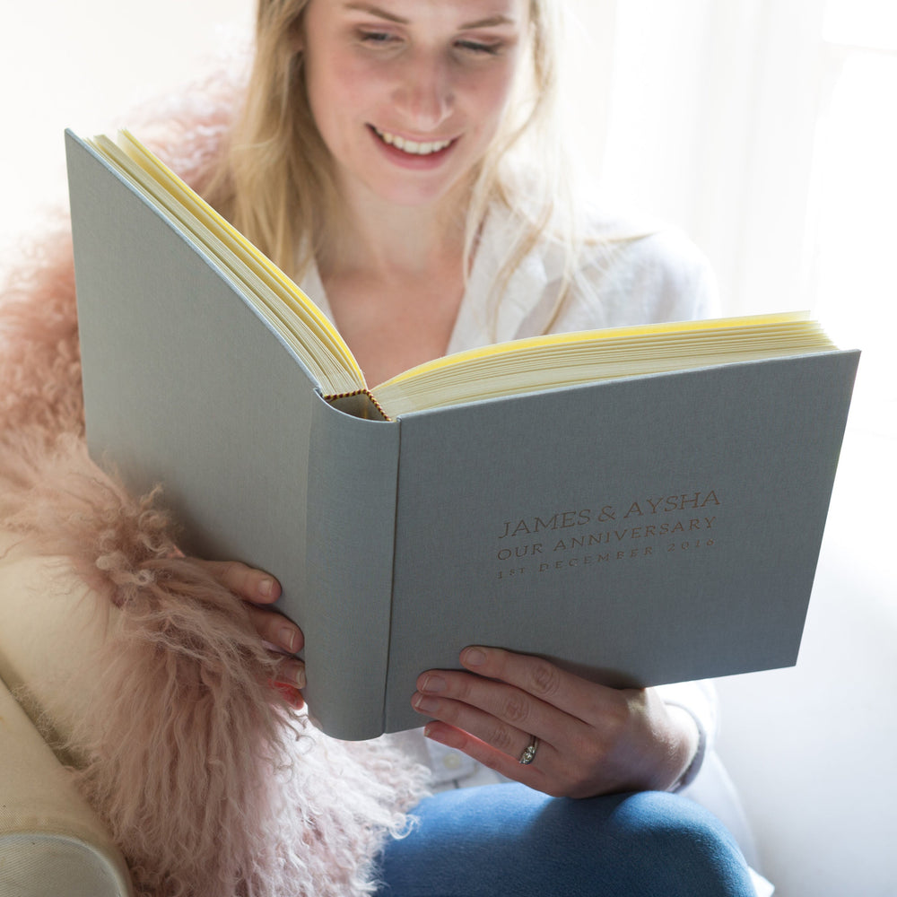 a woman is sitting in a chair, looking through a wedding anniversary album and smiling.