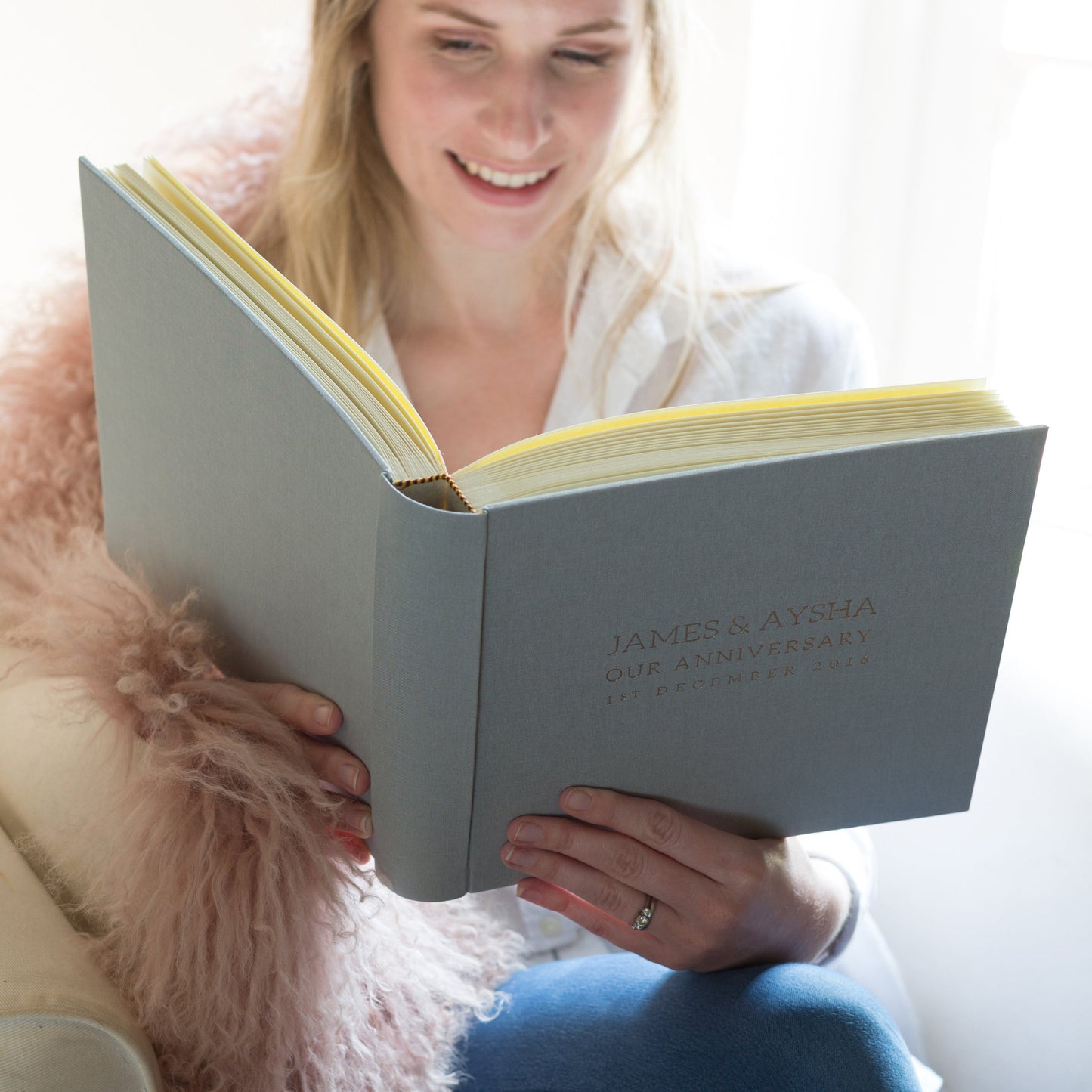 a woman is sitting in a chair, looking through a wedding anniversary album and smiling.