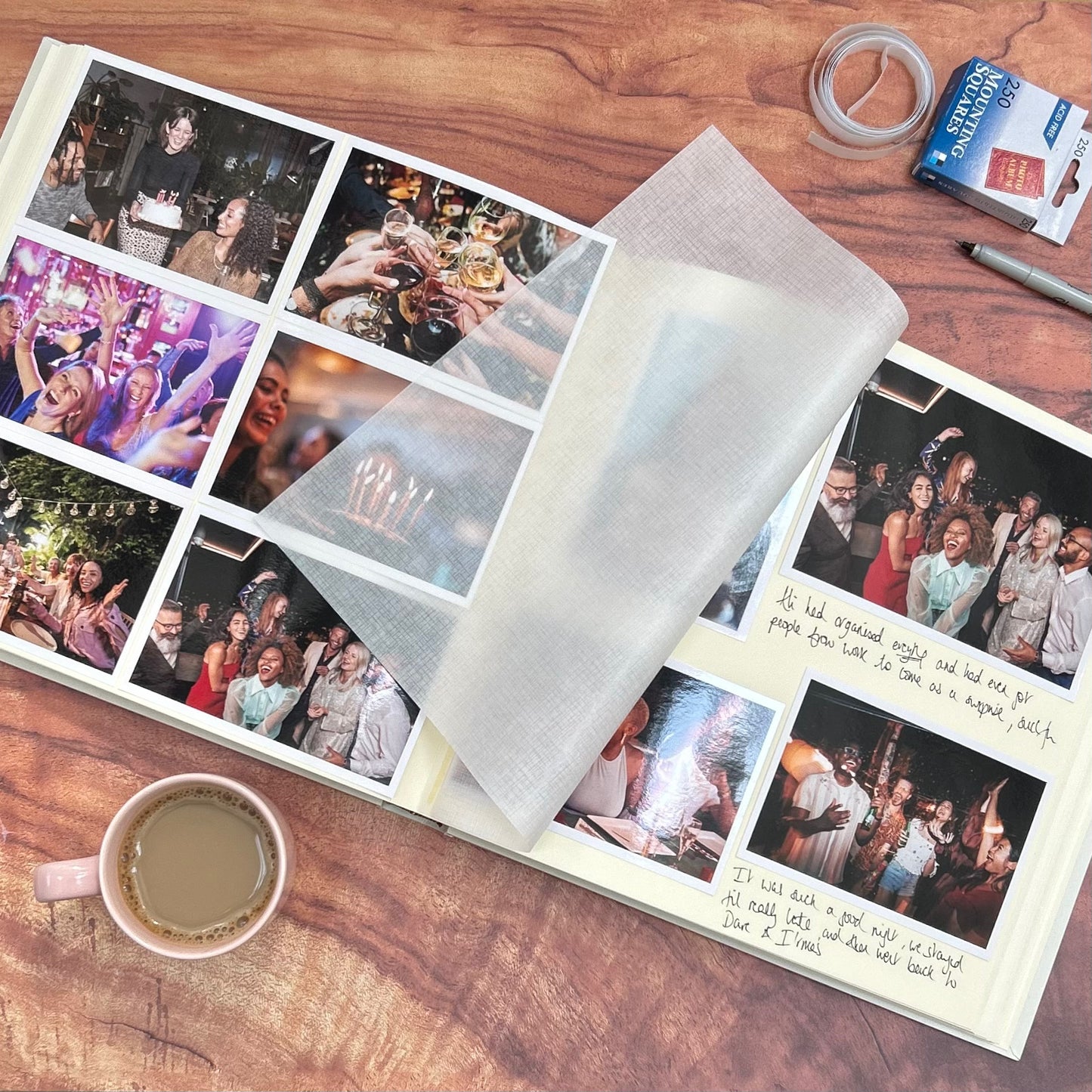 There is a large family album on a wooden table, it has been filled with photographs capturing special memories. There is also a coffee and mounting squares in the shot. 