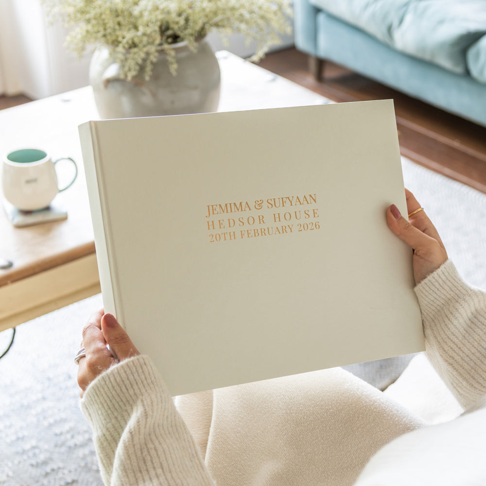 A woman in white skirt and a white jumper is holding a large ivory wedding guest book which has been personalised with gold lettering. There are some flowers in the background.