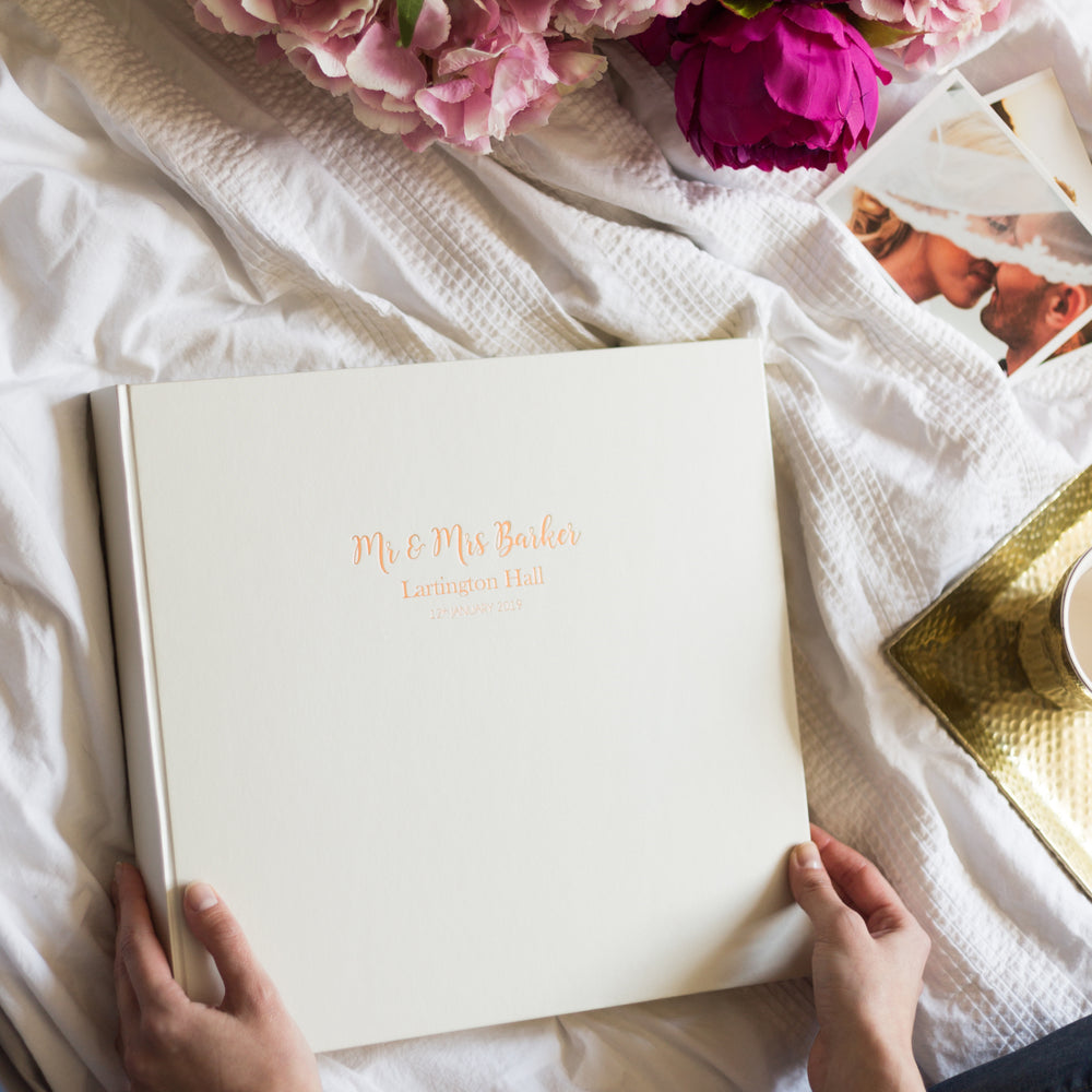 
                      
                        There is a large wedding photo album placed on a bed. A woman is about to open her and her husbands album to place some photos inside. 
                      
                    