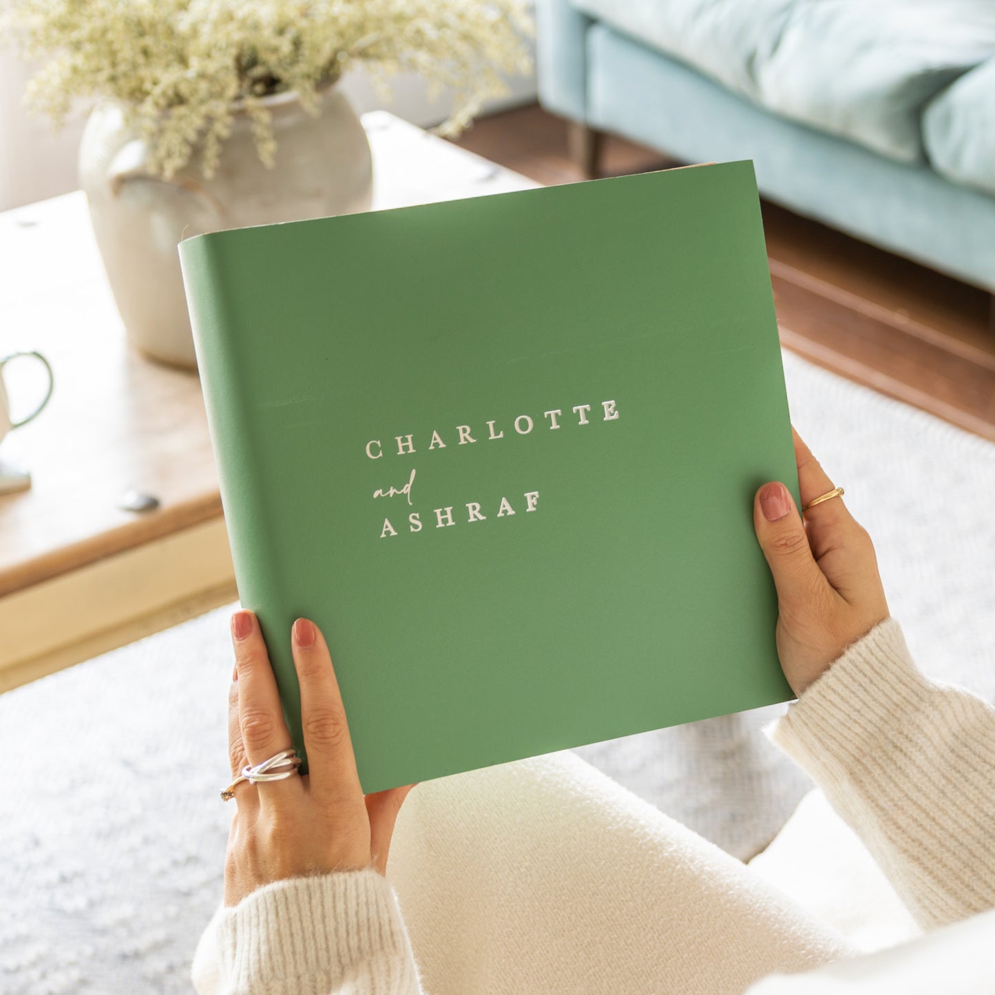 Person holding a green leather photo album in her hands in a cosy home setting. The photo album has been personalised on the front.