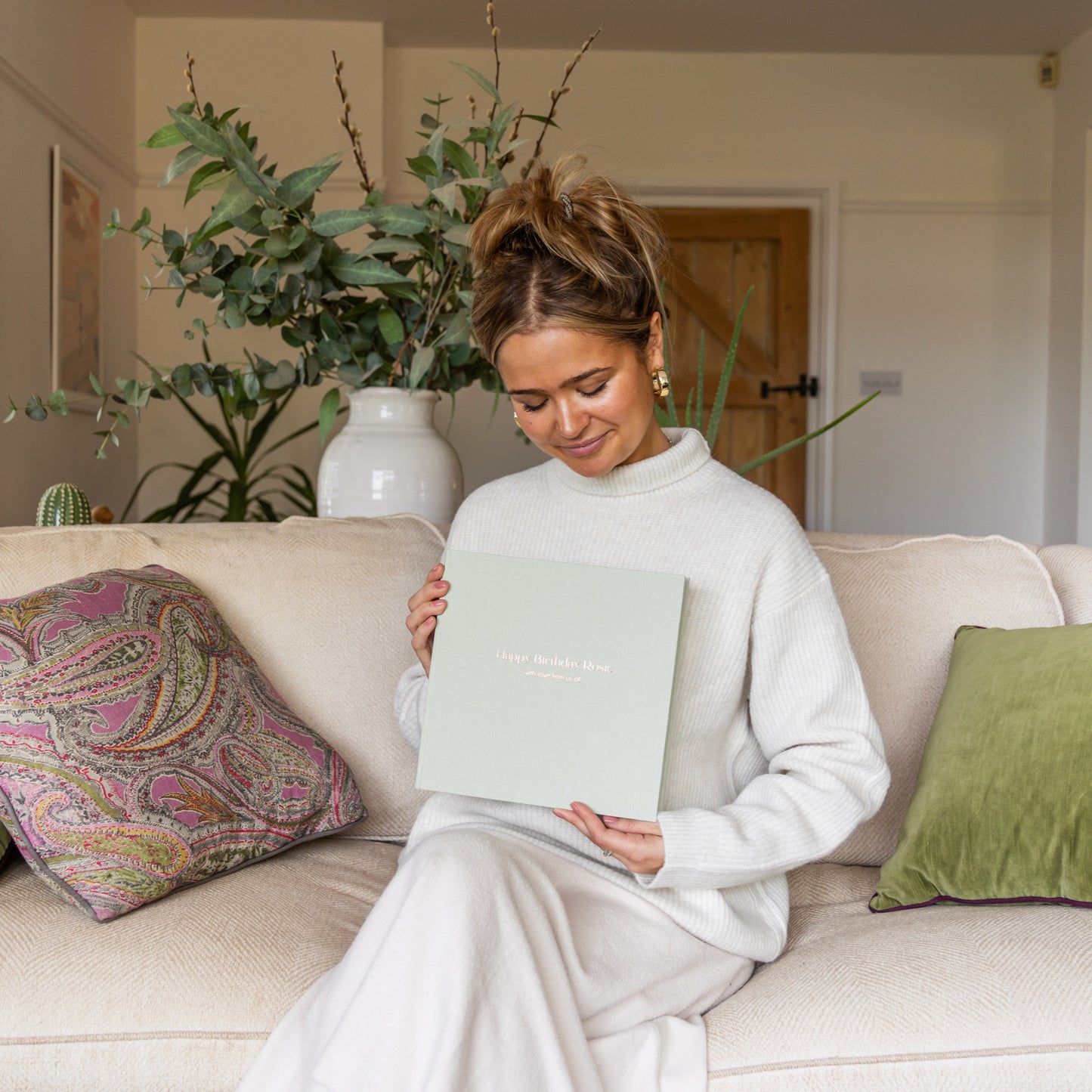Woman sitting on a couch holding a personalised pale green linen-bound photo album  in a cozy living room.