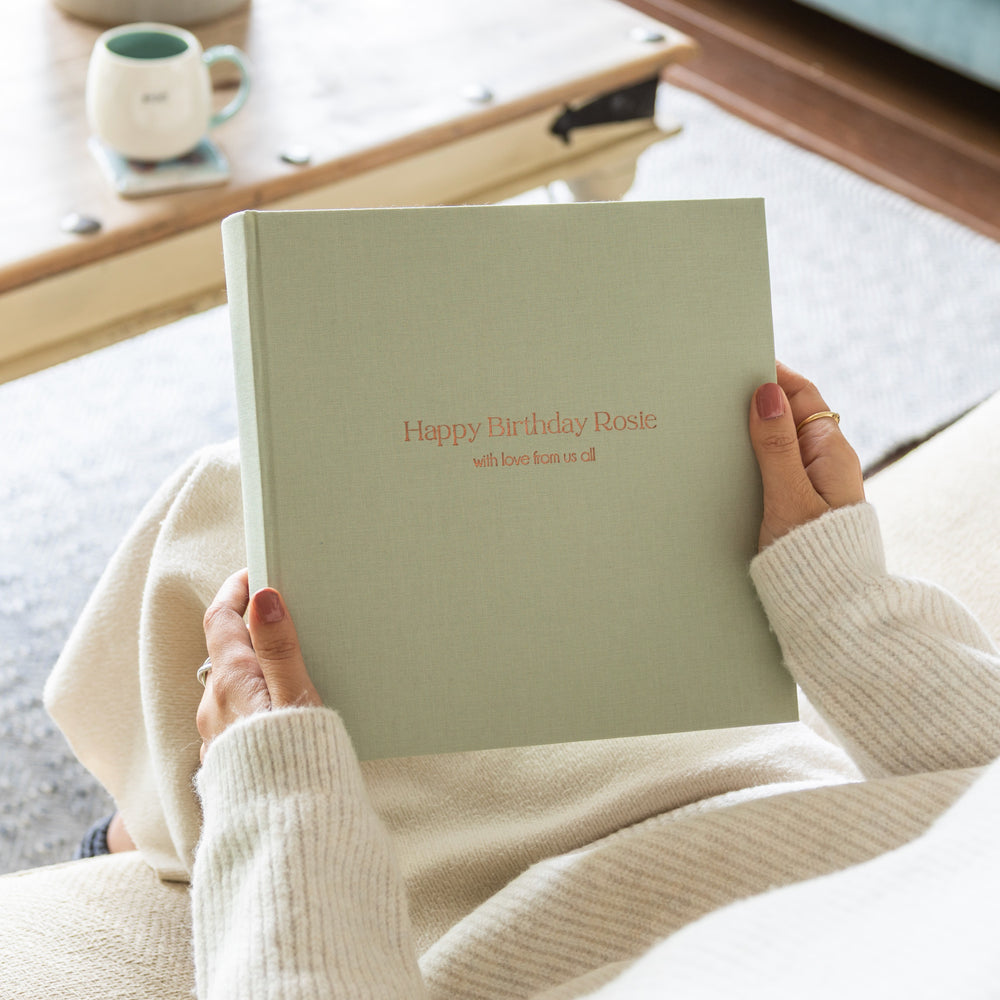 Person holding a personalised pale green linen photo album with 'Happy Birthday Rosie' text, sitting on a couch.