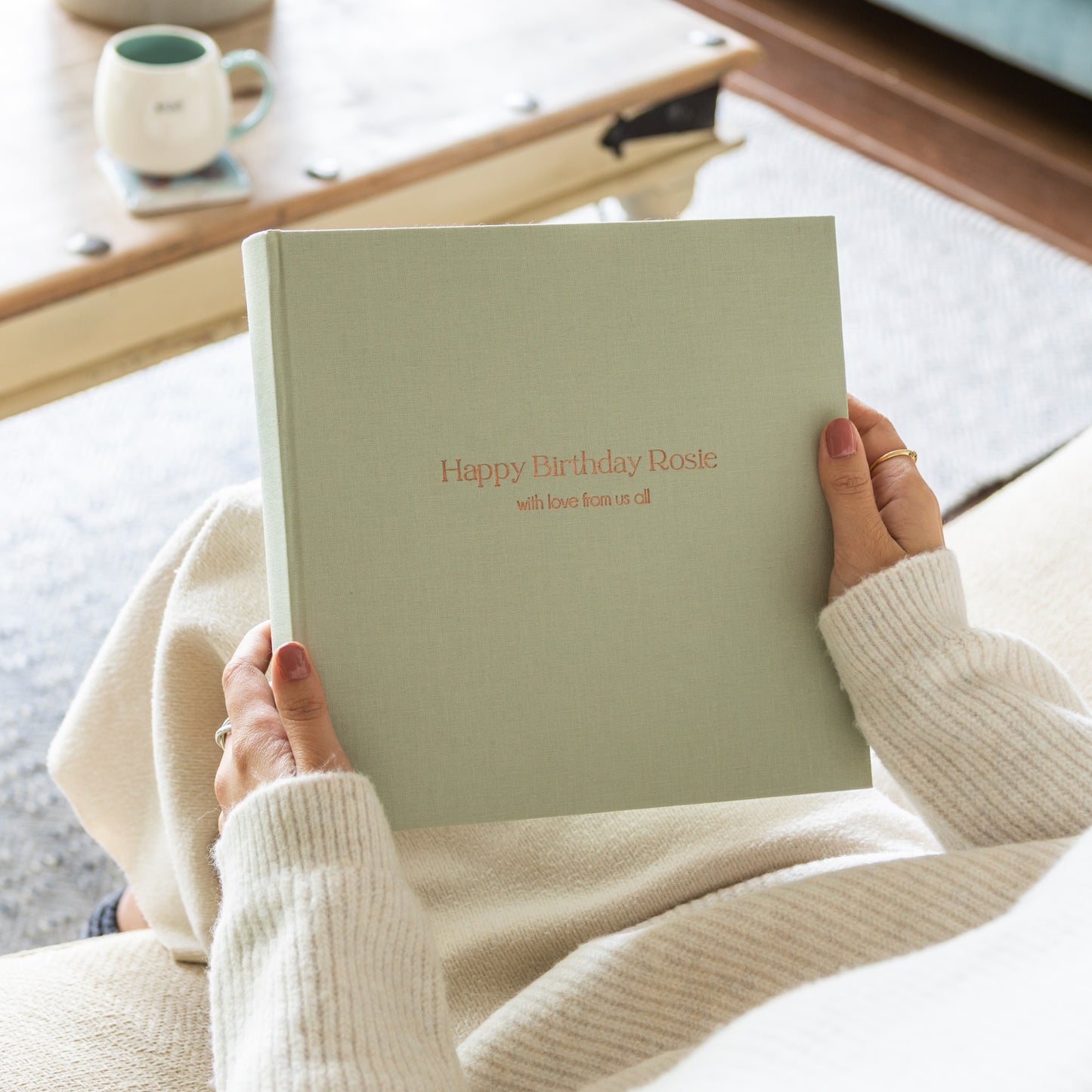 Person holding a personalised pale green linen photo album with 'Happy Birthday Rosie' text, sitting on a couch.