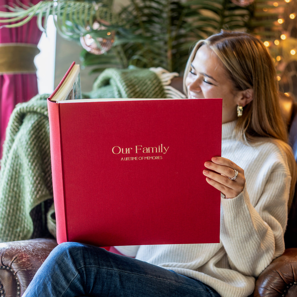 
                      
                        Woman sitting in a chair looking at a red linen personalised photo album printed with 'Our Family'
                      
                    