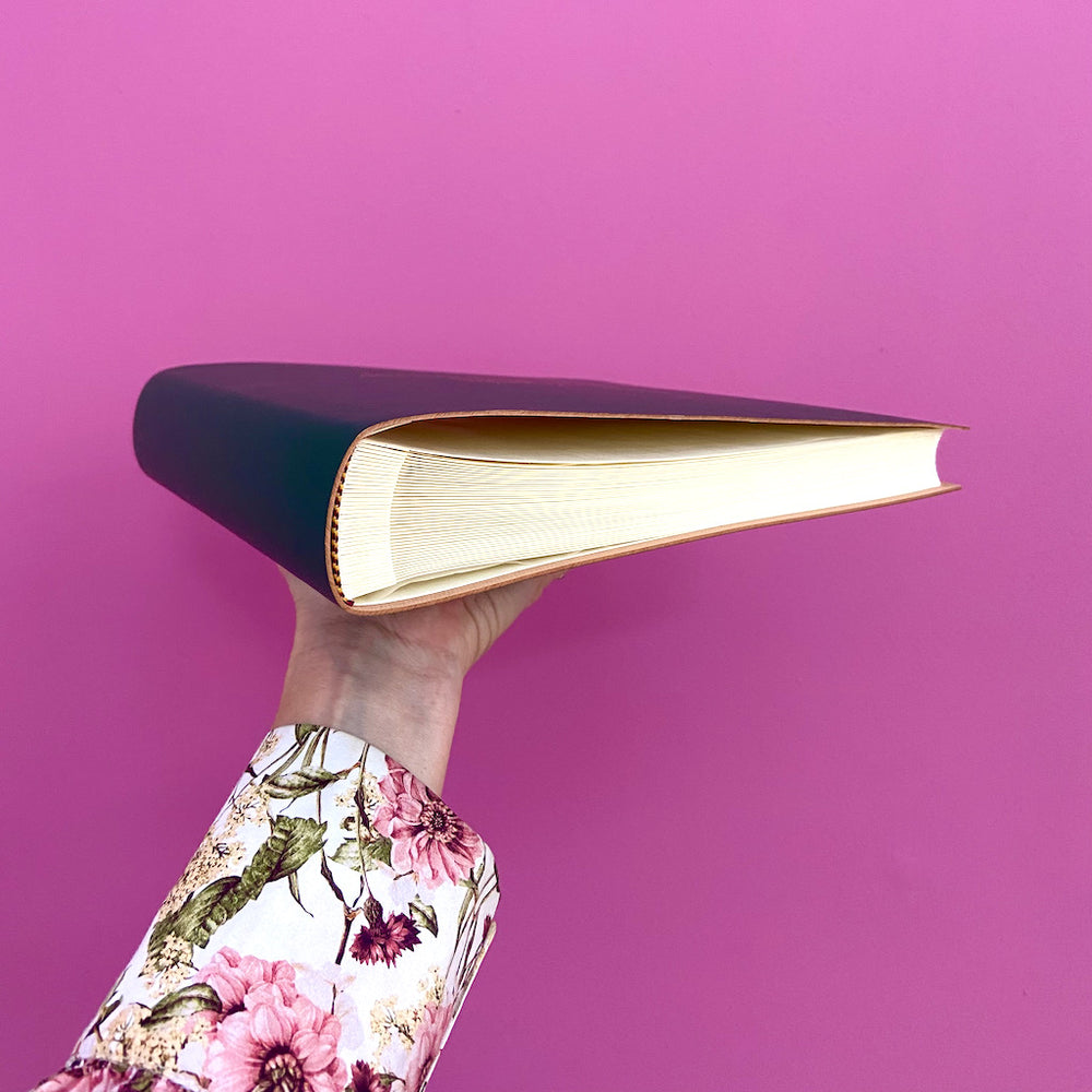 
                      
                        Against a pink backdrop a woman holds a navy blue photo album. You can see the beautifully crafted pages inside.
                      
                    