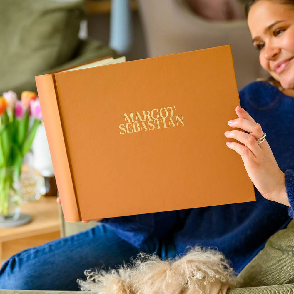 
                      
                        a woman in a blue jumper and a pair of jeans is holding a brown wedding guest book in her hands. The wedding guest book has been personalised on the front with two names in gold writing. In the background there is a vase of flowers.
                      
                    