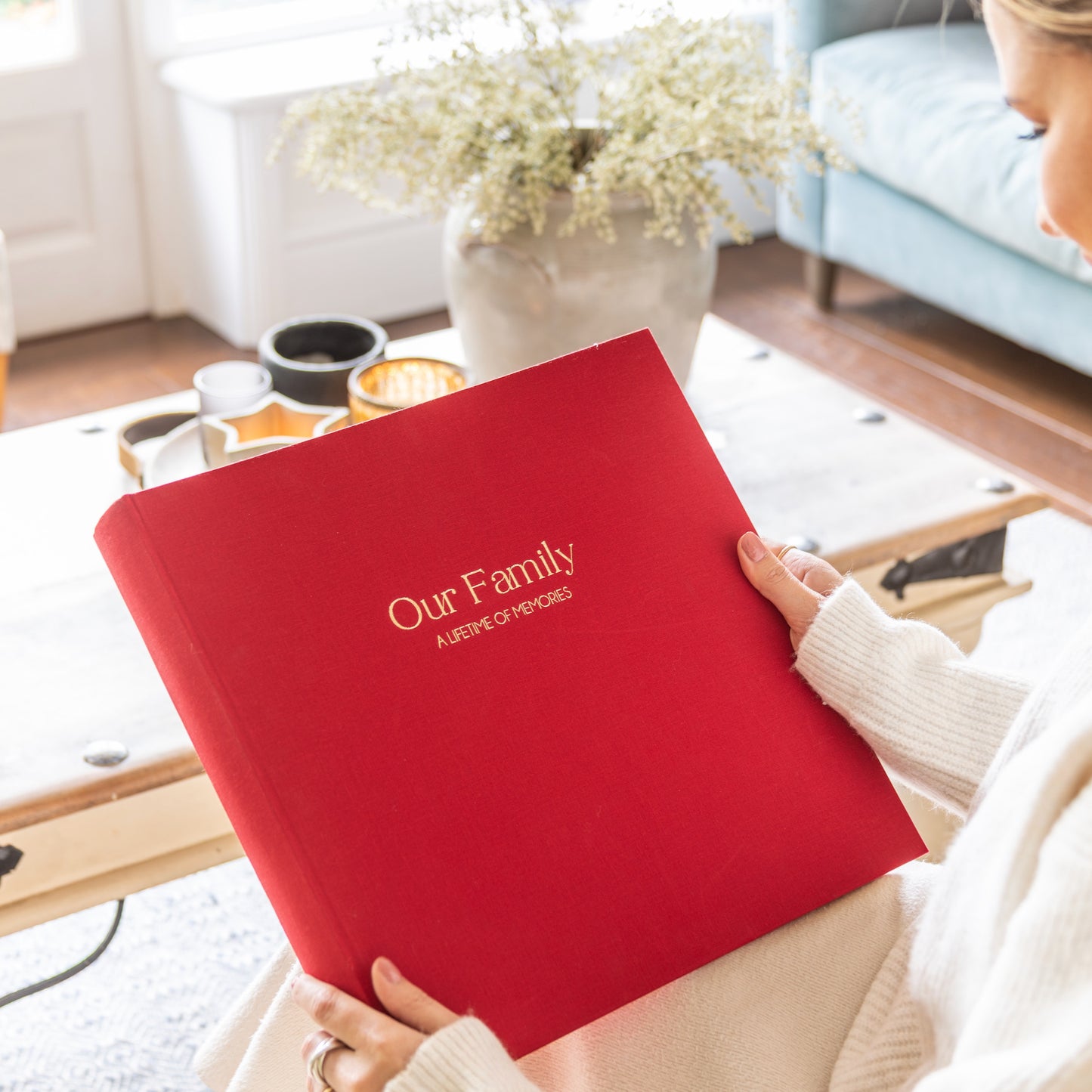 Person holding a red linen personalised photo album titled 'Our Family: A Lifetime of Memories' in a cozy living room.