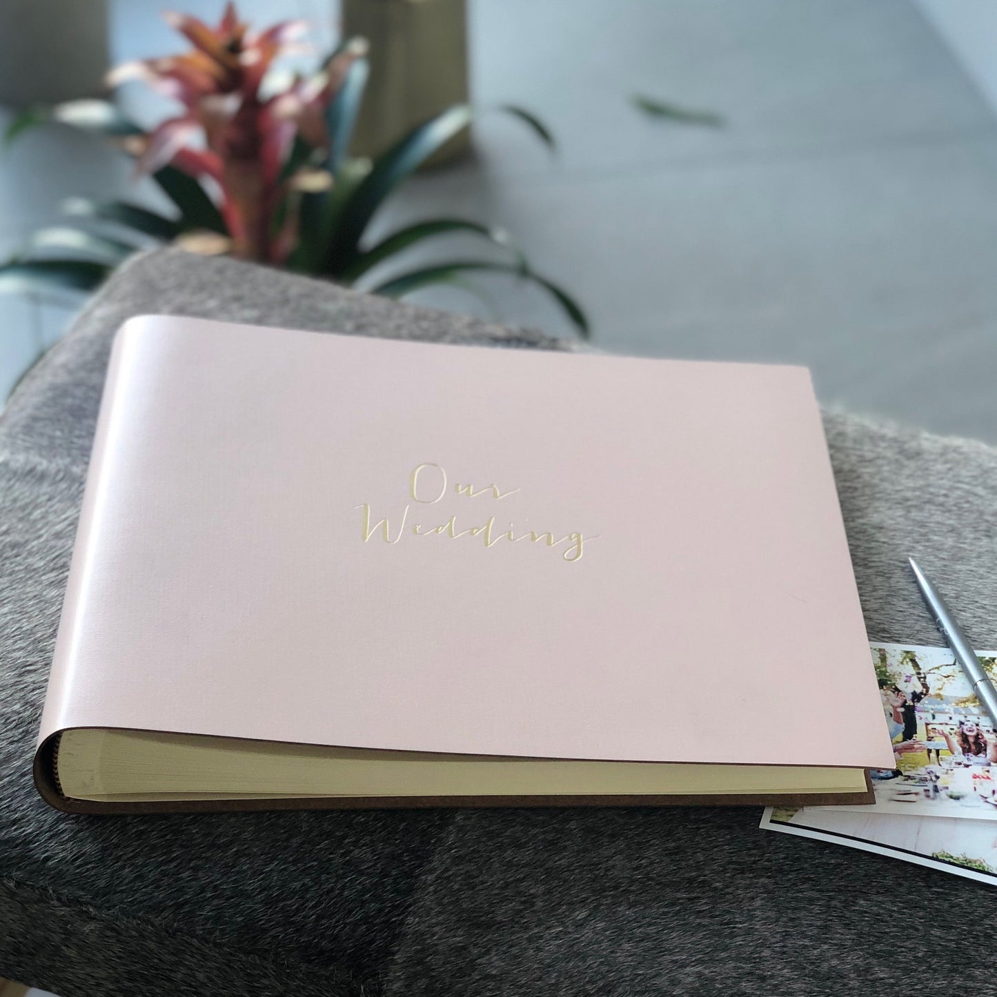 This is a side view of an Our Wedding album placed on a small table. There are photos surrounding the album ready to be placed inside. There is also a pen next to the album to note down memories. 