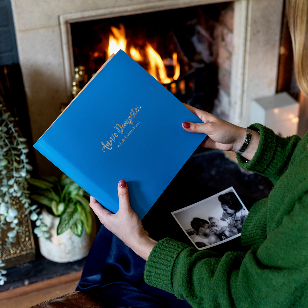 There is a woman wearing a forest green jumper holding a blue condolence book. It has been personalised with silver hot foiling. An old photo is on her lap ready to be placed inside the album.