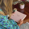 a woman flicks through a pale pink large guest book and then puts it onto a wooden table