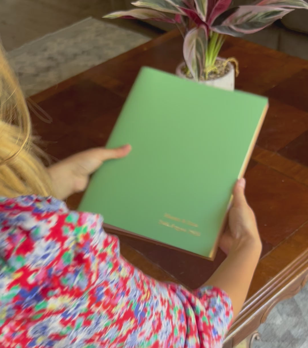A video of a young woman flicking through a green A4 photo album that has been personalised on the front. She places it on a coffee table where there is a house plant. 