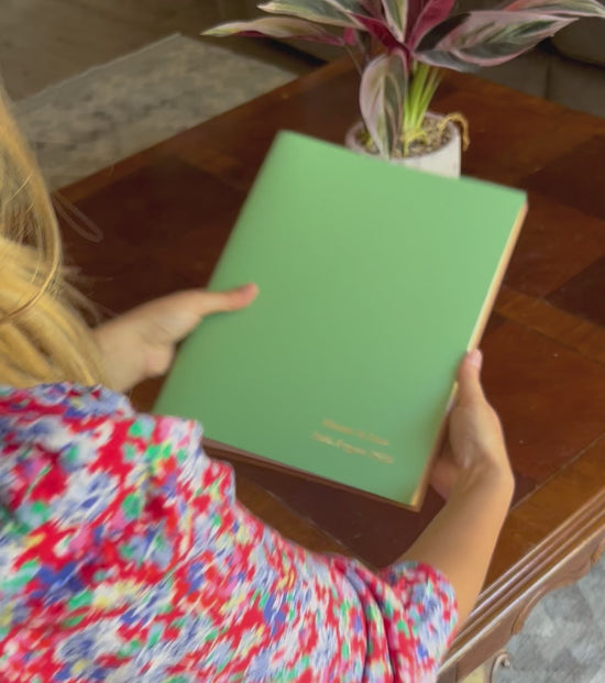 Load and play video in Gallery viewer, a video of a young woman flicking through a green A4 photo album that has been personalised on the front. She places it on a coffee table with a plant on it
