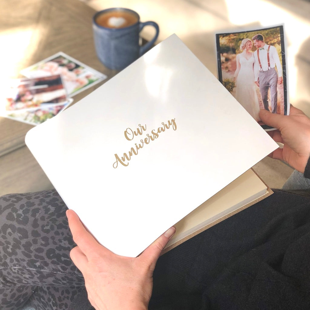a white leather photo album is being held on a woman's lap. The photo album is printed with the words Our Anniversary on the front