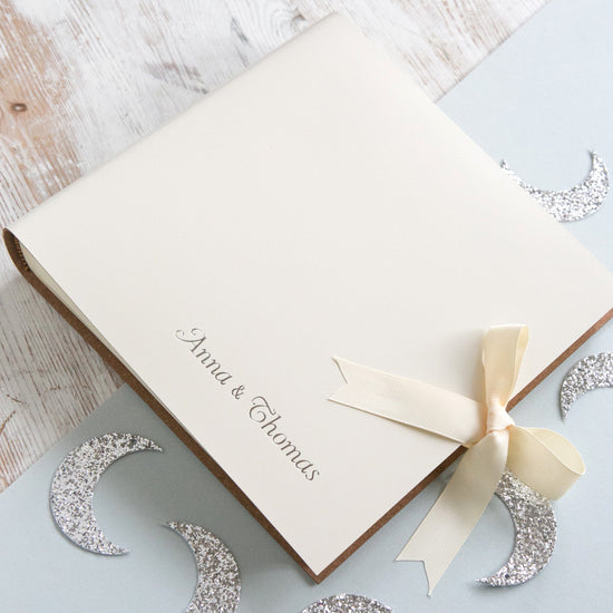 Load image into Gallery viewer, you can see the ivory baby album on a table. It has been printed on the front with a couple of names in silver writing. The ribbon has been done up in a bow
