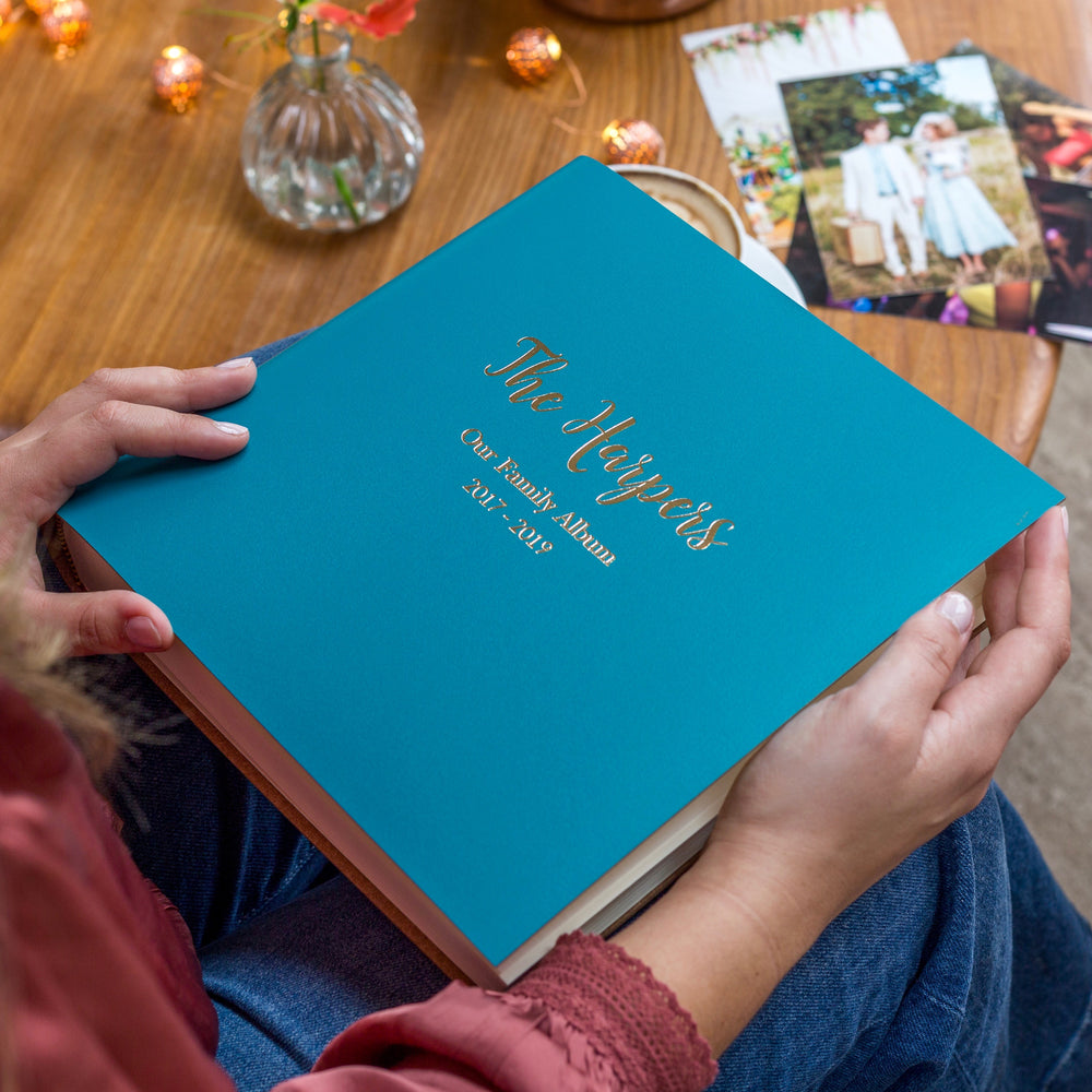 a square photo album is resting on someone's knee. It has been personalised. There are photos on the table