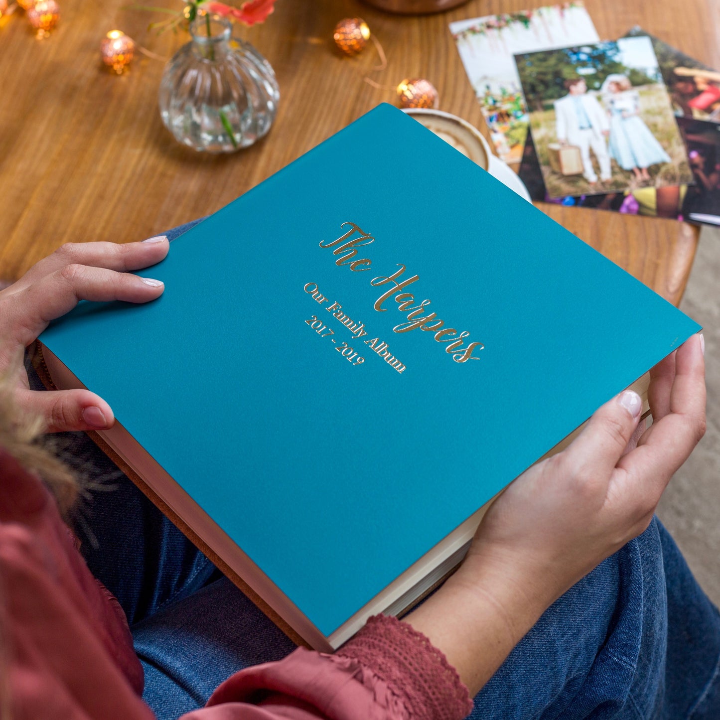 a square photo album is resting on someone's knee. It has been personalised. There are photos on the table
