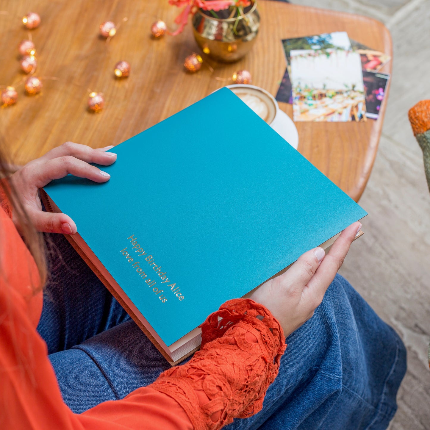 a square photo album is resting on someone's knee. It has been personalised. There are photos on the table