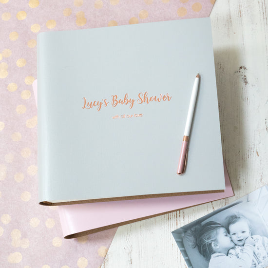 a baby shower photo album is on a table and there is a pen on top of the  album. There is also a photo of a small child with a baby next to the photo album