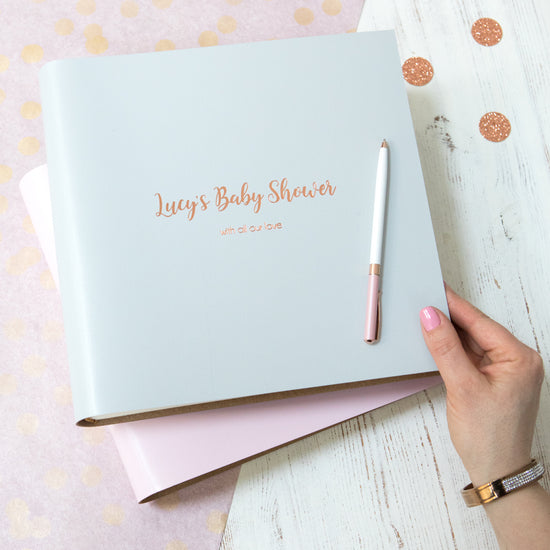 a pale grey baby shower photo album is on a table and is surrounded by little rose gold dots. The baby shower photo album has been printed with someone's name. 