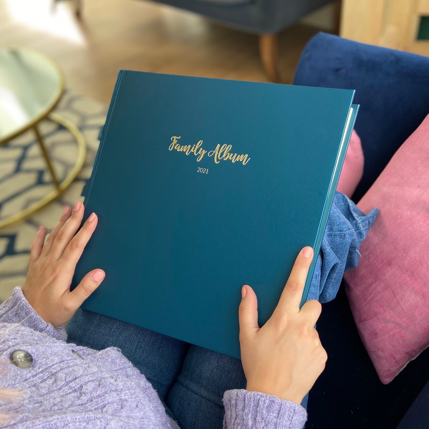 A woman is sitting on a sofa with her knees up. A blue family album is in her lap and she has her hands on the photo album. The album has been printed with gold hot foil. 