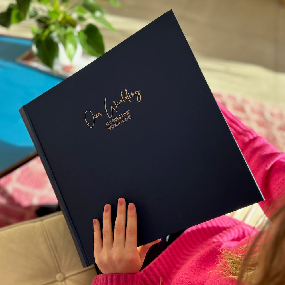 
                      
                        A lady in a pink jumper looking through a navy blue photograph album with "Our Wedding" embossed in gold on the front
                      
                    