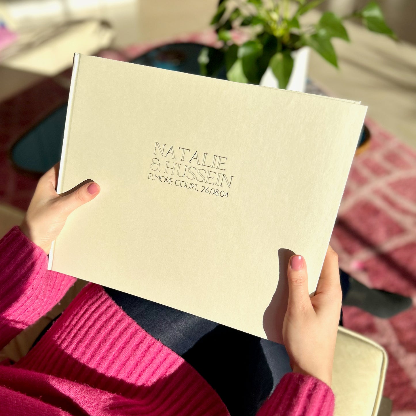Load image into Gallery viewer, an ivory wedding guest book is being held by a woman sitting on the sofa. The sun is reflecting off the guest book showing its iridescent bookcloth
