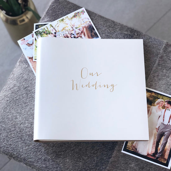 a square wedding album lies on a grey stool surrounded by wedding photos. The wedding album has been printed with Our Wedding on the front in gold foil