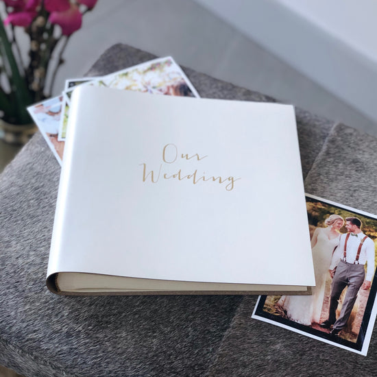 a white square leather photo album lies on a grey stool surrounded by weddng photos. It has been printed with the words Our Wedding on the front