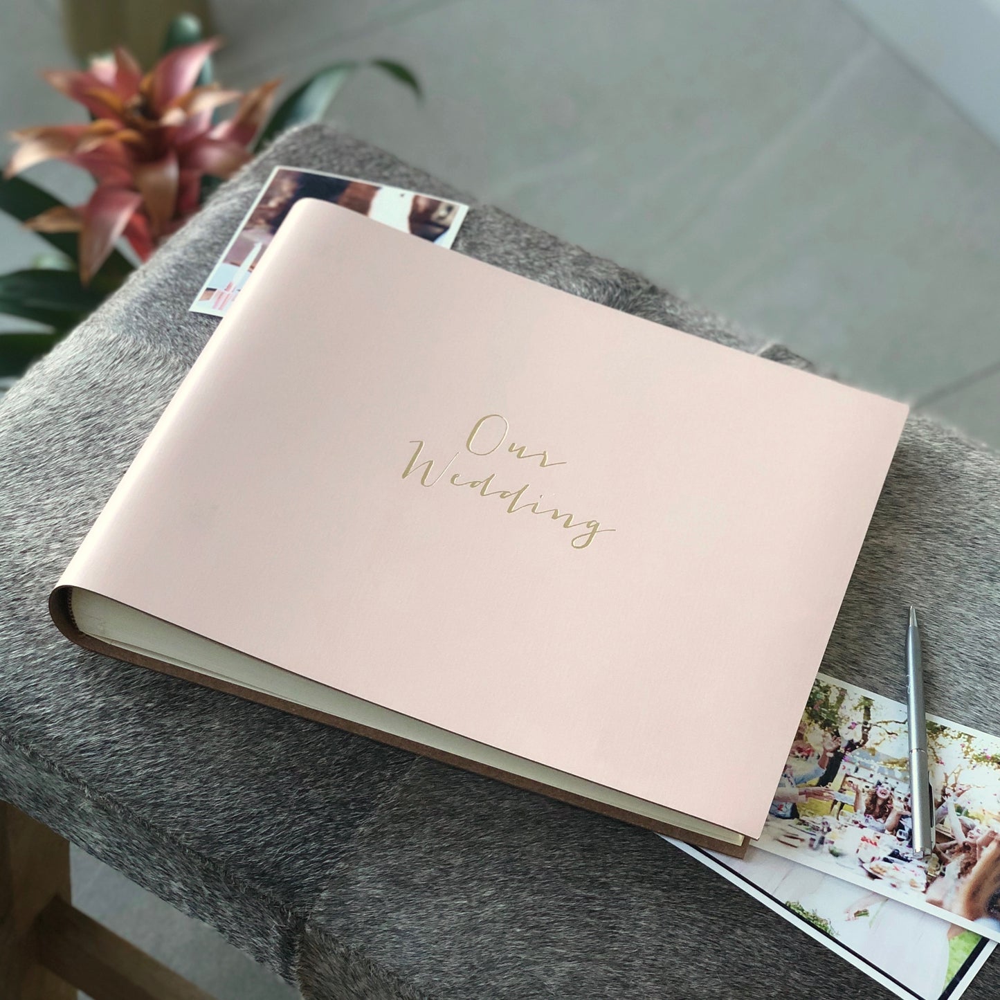 on a grey footstool is a pale pink wedding guest book, It has been pritend with the words Our Weddingon the front . There are wedding photos and a pen also on the stool
