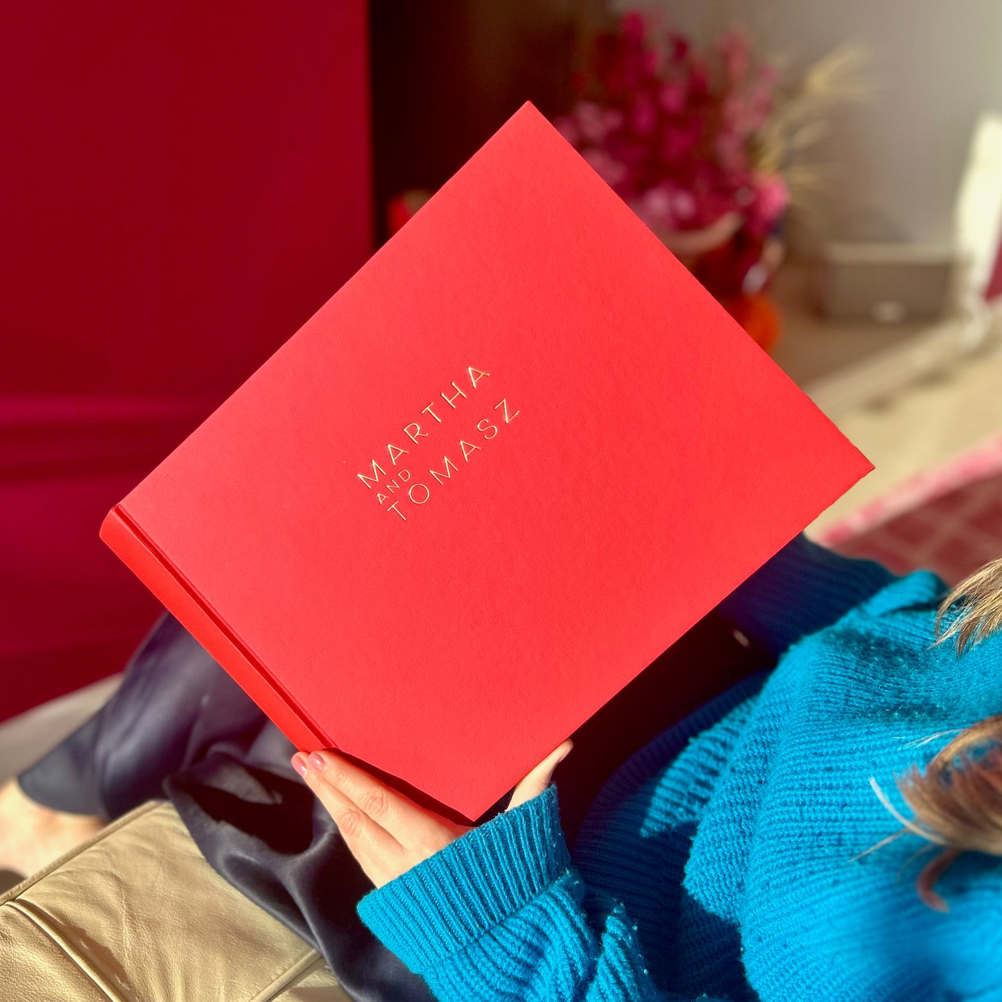 a woman is sitting on the sofa looking through a big red wedding guest book. It has gold writing on the front