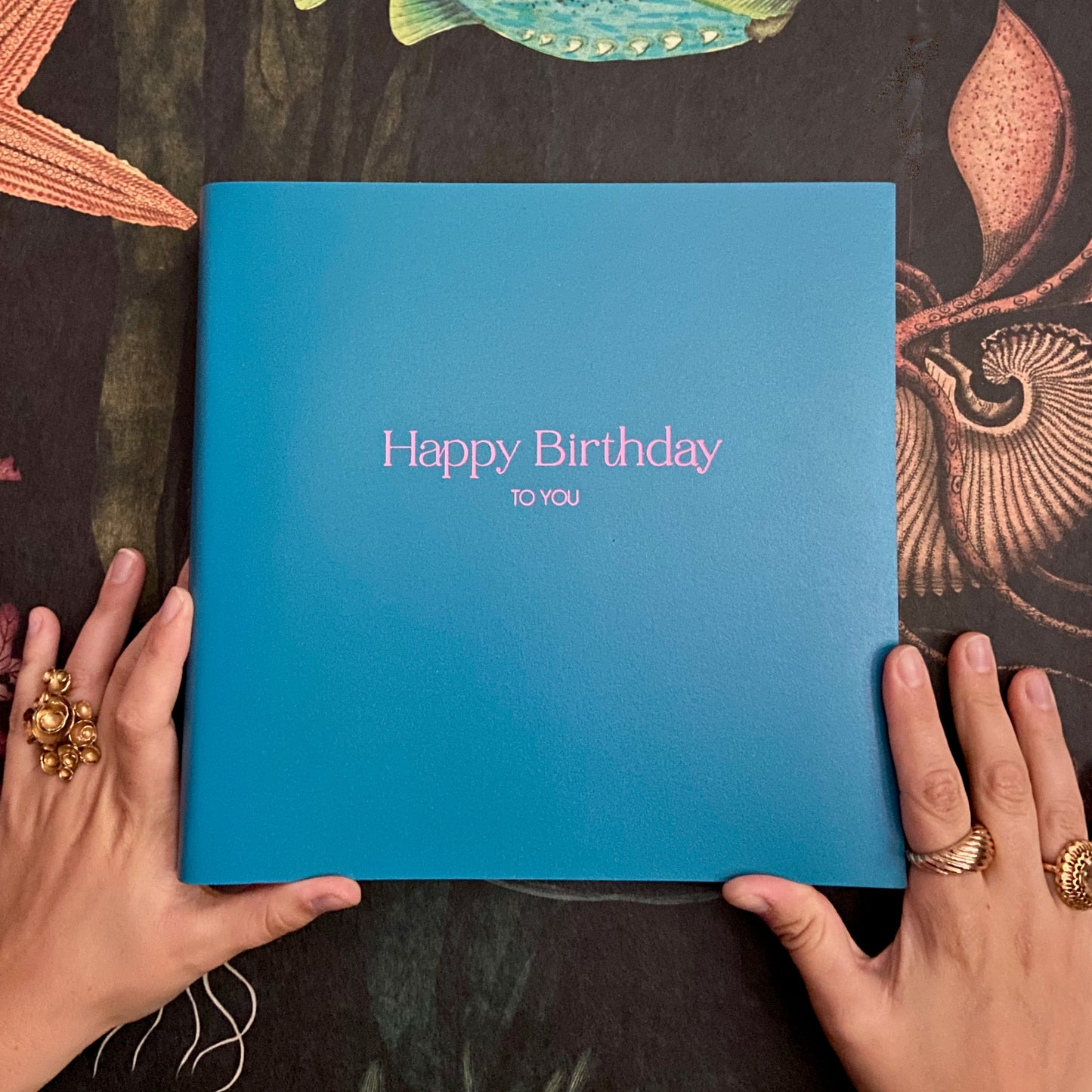 against a black patterned back drop there is a blue photo album which has been printed with the words Happy Birthday to you. The birthday phtoo album is blue and the writing on the front of the album is pink