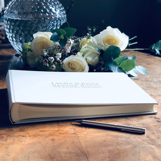 on a table is a wedding guest book next to some flowers. It is ready for the wedding guests to sign and write their messages