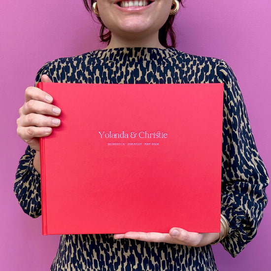 a woman in an animal print dress is standing in front of a pink wall. She is holding a red wedding guest book that has been personalised in a pink foil