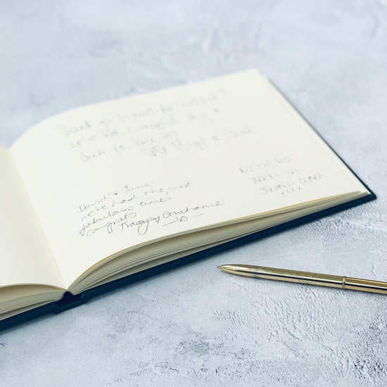 Load image into Gallery viewer, a wedding guest book lies open on a table and you can see the messages which have been written inside it. there is a pen next to the guest book
