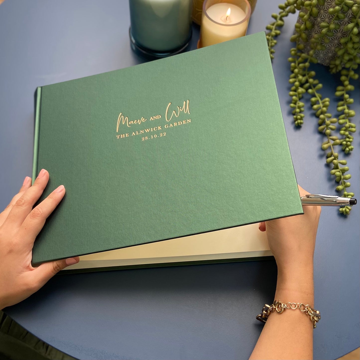 A beautiful green wedding guest book resting on a blue table with candles and potted plants in the background