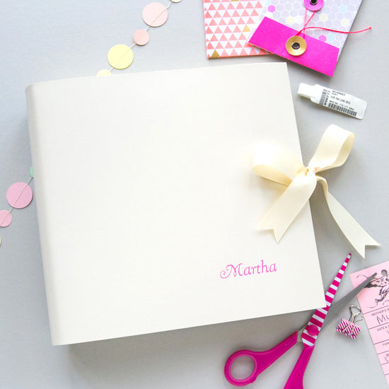 a white leather baby memory book lies on a grey background. The memory biook has been printed with the name Martha on the front in oink writing. There are a pair of scissors also on the table 