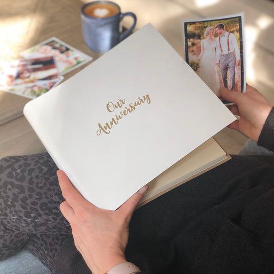 a white wedding anniversary photo album is being held by a woman. There is a cup of coffee in the bacj ground