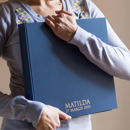 a large navy blue photo album is being held by a woman in a blue jumper. There is writing on the bottom of the album in gold foil