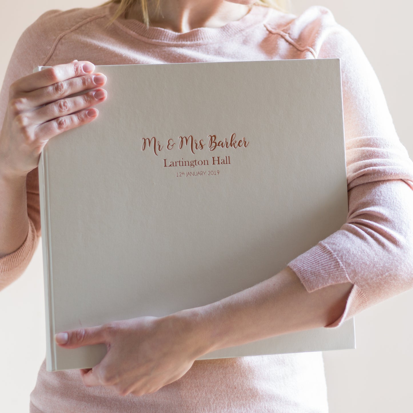a girl in a pink jumper is clutching a large ivory wedding photo album. The photo album has wedding details hotfoil printed onto the front