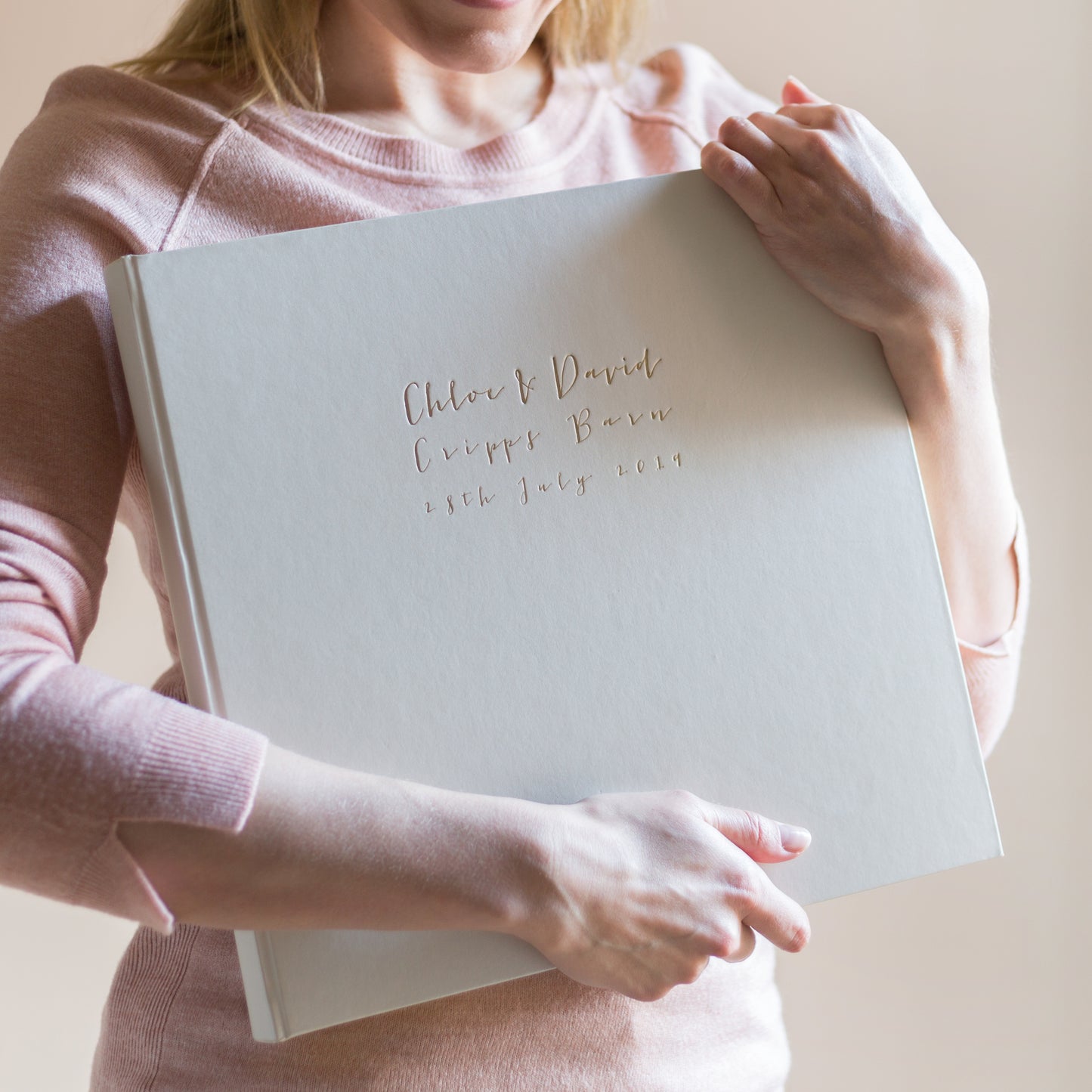 a woman in pink top holds a large ivory wedding album that has been printed on the front with gold foil 
