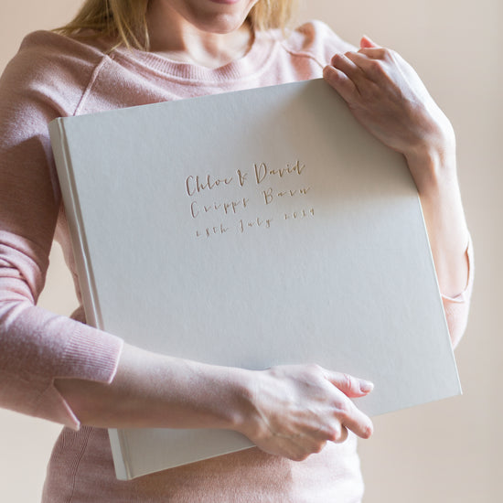 a woman in pink top holds a large ivory wedding album that has been printed on the front with gold foil 