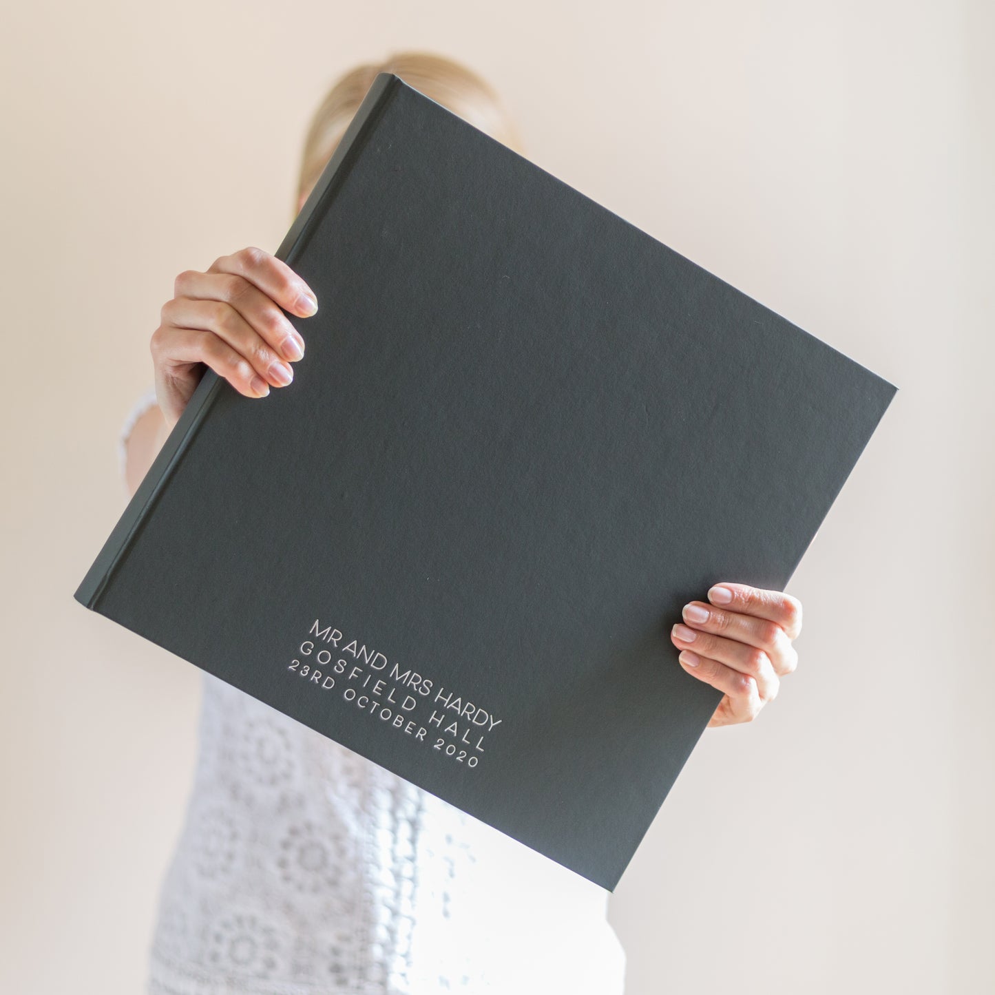 a woman is holding a large black wedding photo album in outstretched arms. The wedding album has been printed on the bottom with the bride and grooms wedding details 
